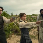 "Three people in vintage clothing stand outdoors in a rugged, green landscape; two point pistols ahead while the third stands beside them with a cane, all looking alert and tense."