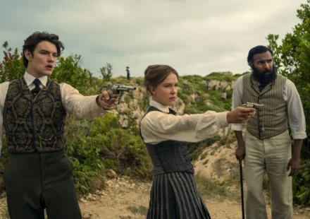 "Three people in vintage clothing stand outdoors in a rugged, green landscape; two point pistols ahead while the third stands beside them with a cane, all looking alert and tense."