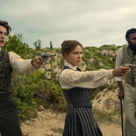 "Three people in vintage clothing stand outdoors in a rugged, green landscape; two point pistols ahead while the third stands beside them with a cane, all looking alert and tense."