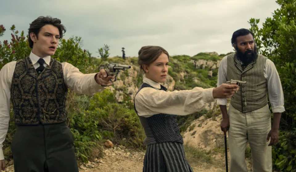 "Three people in vintage clothing stand outdoors in a rugged, green landscape; two point pistols ahead while the third stands beside them with a cane, all looking alert and tense."