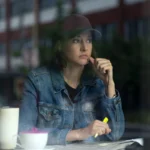 "Woman in denim jacket and baseball cap sits at a table inside a cafe, looking thoughtful while holding a yellow highlighter, with a mug and papers in front of her, city street visible through the window."