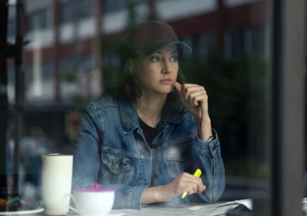 "Woman in denim jacket and baseball cap sits at a table inside a cafe, looking thoughtful while holding a yellow highlighter, with a mug and papers in front of her, city street visible through the window."