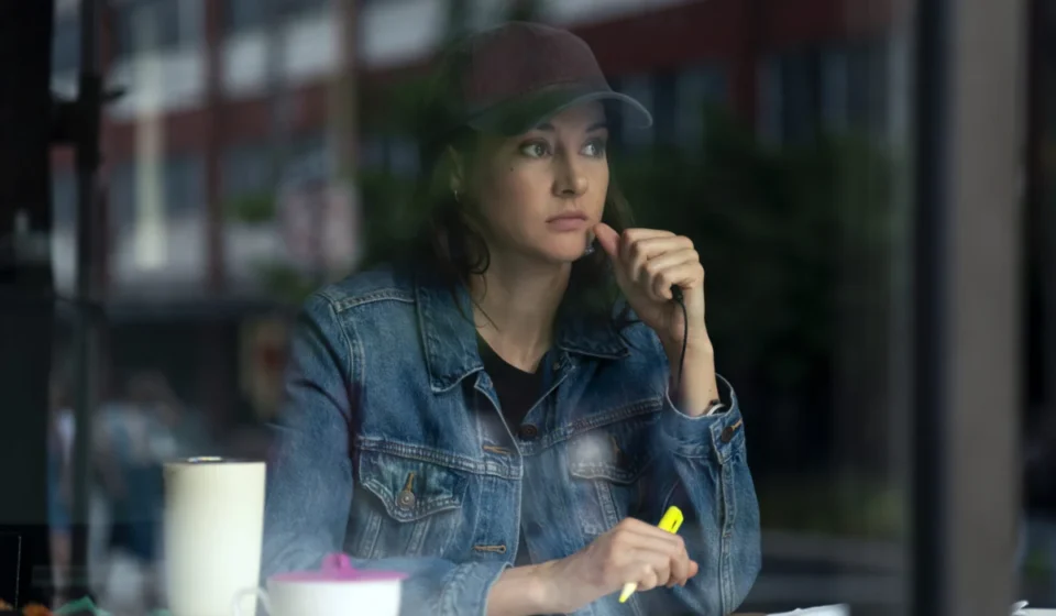 "Woman in denim jacket and baseball cap sits at a table inside a cafe, looking thoughtful while holding a yellow highlighter, with a mug and papers in front of her, city street visible through the window."
